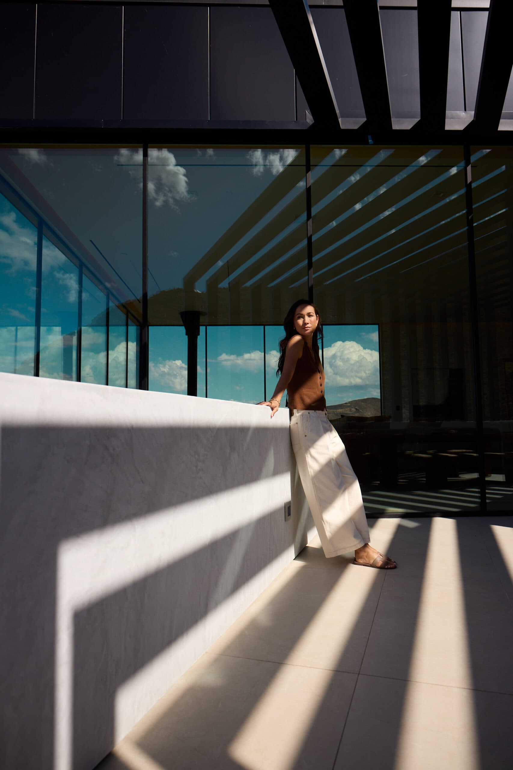 Woman standing on a modern balcony with glass walls and architectural shadows, overlooking mountain views under a bright blue sky.