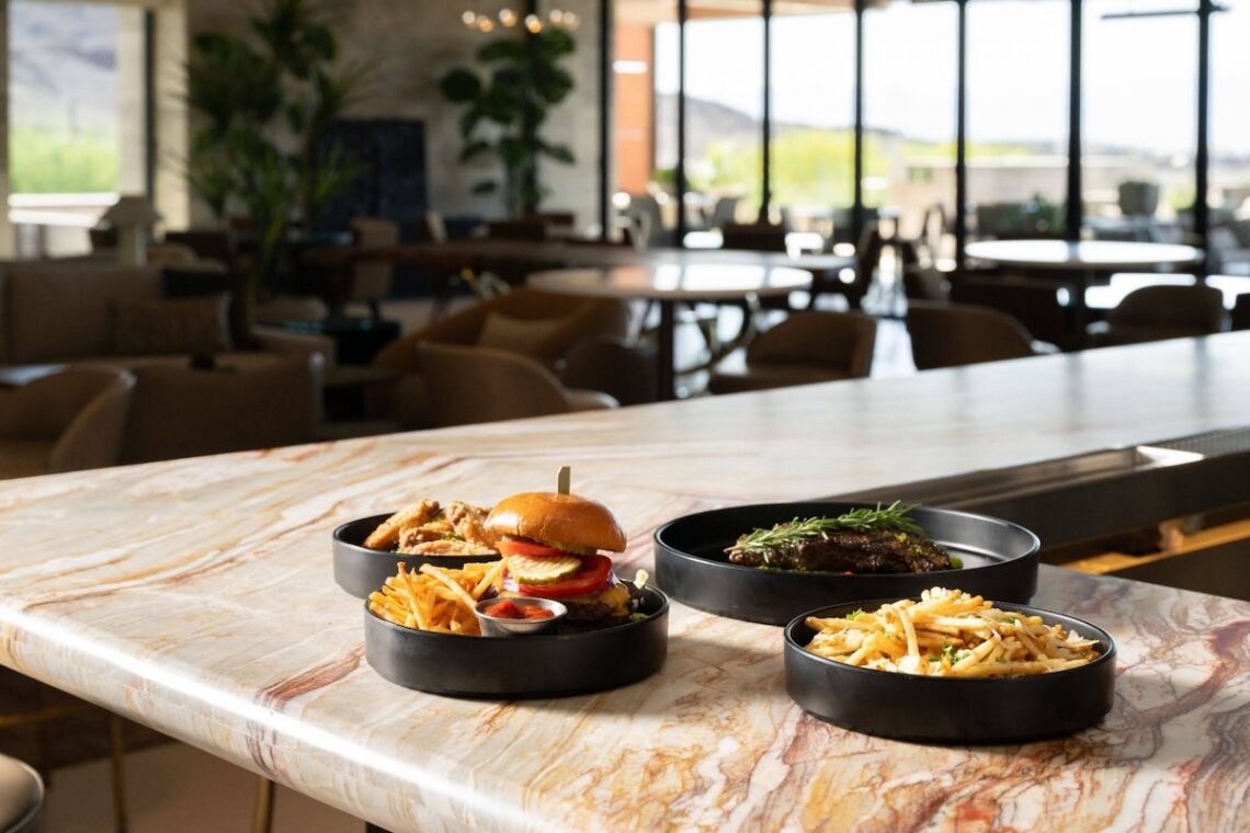 Plated restaurant dishes on a marble counter, including a burger with fries, steak, and pasta, set in a modern dining space with large windows and natural light.