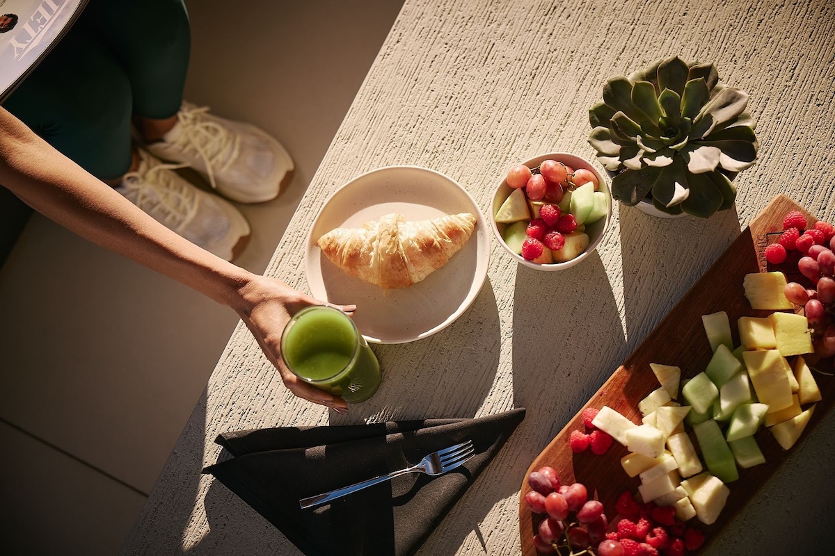 Top-down view of a light-filled table with a croissant on a plate, a green smoothie in hand, a bowl of mixed fruit, and a charcuterie-style board with cheese and fruit beside a small potted succulent.