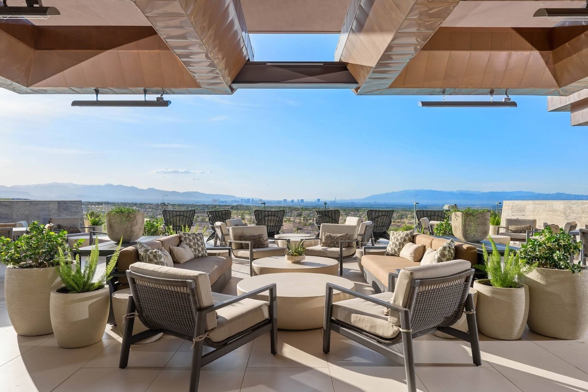 An upscale outdoor lounge with cushioned seating, plants, and a wide open view of a city skyline and mountains in the distance.