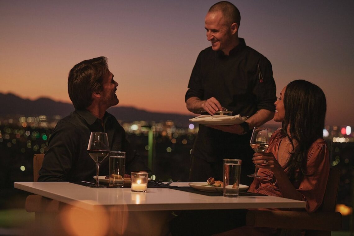 A server presenting a dish to a couple dining at a candlelit table on a rooftop at sunset, with a glowing city skyline in the background.
