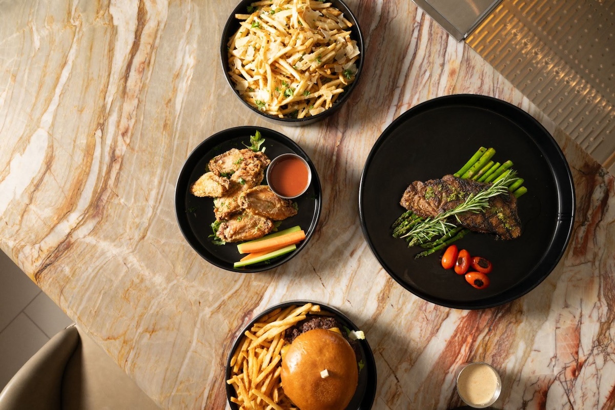 Overhead view of a restaurant table with plated dishes including a burger and fries, fried chicken wings with dipping sauce, a steak with asparagus and cherry tomatoes, and a bowl of truffle fries.
